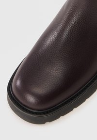 Stivale in pelle viola scuro con una superficie liscia, punta arrotondata e suola in gomma nera con cuciture decorative lungo il bordo.