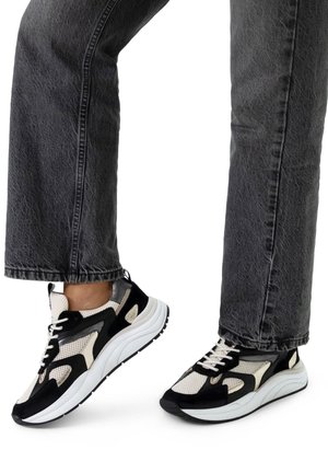 Black and white athletic sneakers with mesh and suede materials, featuring a chunky sole and contrasting accents, paired with gray jeans.