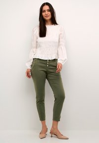 Bluză albă cu pliuri și mâneci bufante, pantaloni în nuanța verde măsliniu, cu croială conică și detalii cu butoane, și sandale cu toc în nuanța nud. Design simplu și elegant.