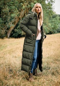 Cappotto lungo, verde oliva, imbottito, con chiusura a zip e tasche laterali, indossato sopra un maglione chiaro e jeans blu, abbinato a stivaletti marroni.