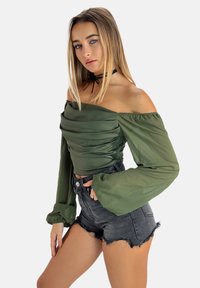 Blusa verde off-the-shoulder con frontale arricciato, maniche lunghe a sbuffo in tessuto trasparente, abbinata a pantaloni corti in denim neri a vita alta con orlo sfilacciato.
