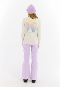 Beige top met lange mouwen en een kleurrijke bergafbeelding; gecombineerd met flare-lavendelbroeken en een lavendelbeanie. Zwarte laarzen maken de look compleet.