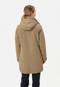 Donna con capelli biondi raccolti in una coda di cavallo, che indossa un cappotto con cappuccio color cammello e pantaloni neri, girata di spalle su uno sfondo bianco.