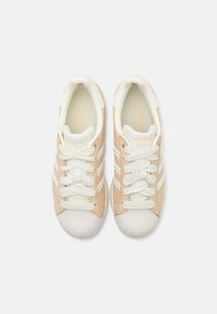 Baskets beiges avec des accents blancs et trois rayures. Elles présentent un capuchon en caoutchouc texturé blanc et des lacets tissés. Tige en synthétique lisse.