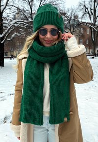 Zielona, prążkowana czapka i pasujący szalik, założone na kremowy sweter i beżowy płaszcz z kołnierzem z jagnięcej wełny, na tle zaśnieżonego krajobrazu.