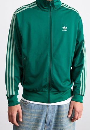 Person, die eine grüne Adidas Trainingsjacke mit weißen Streifen an den Ärmeln und Logo auf der Brust trägt, kombiniert mit hellblauen Jeans.