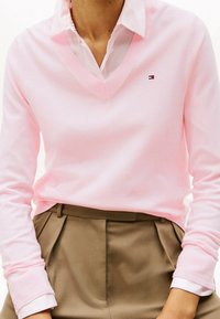 Maglione rosa chiaro a V realizzato in materiale morbido, con polsini e orlo a coste. Logo sul lato sinistro del petto, indossato sopra una camicia bianca con colletto.