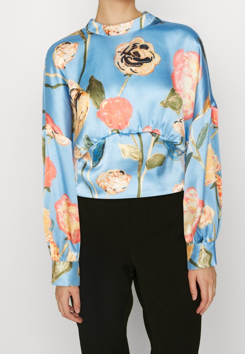 Blusa in satin blu con stampa floreale, caratterizzata da maniche lunghe a sbuffo, un collo alto e una vita arricciata; abbinata a pantaloni neri.