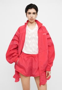 Jong persoon draagt een felrode Adidas windbreaker jas en bijpassende korte broek over een wit shirt met V-hals en patroon, tegen een witte achtergrond.