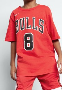 Dítě oblečené v červeném tričku s logem Chicago Bulls s číslem 8 a odpovídajícími červenými šortkami na jednobarevném pozadí.