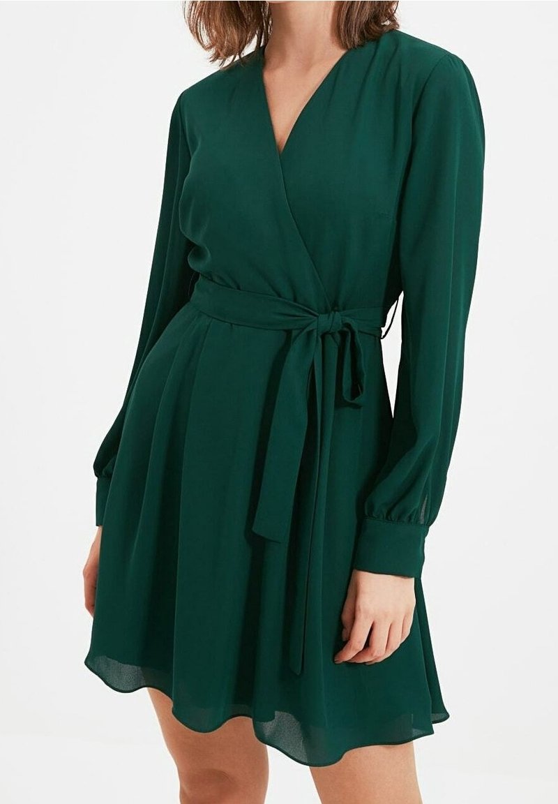 Femme portant une robe portefeuille verte foncée à manches longues avec une ceinture à la taille et une jupe fluide, debout devant un fond blanc uni.
