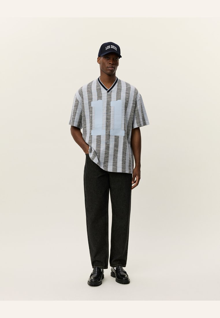 Camisa de manga curta riscada em cinza e branco com o número "11" em azul, combinada com calças escuras e um boné preto com texto branco.