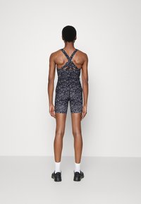 Czarny i szary bodysuit w wzory z krzyżowymi ramiączkami, dopasowany krój i krótkie nogawki. Noszony z czarnymi sneakersami i białymi skarpetkami.