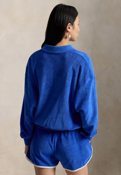 Mujer con cabello largo y oscuro, vestida con una camiseta de manga larga azul y pantalones cortos a juego con ribete blanco, de espaldas sobre un fondo liso beige.