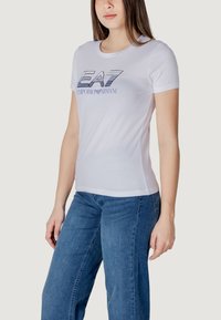 T-shirt bianca a maniche corte con scollatura a girocollo e logo "EA7 Emporio Armani" stampato in blu. Tessuto morbido con una silhouette aderente.