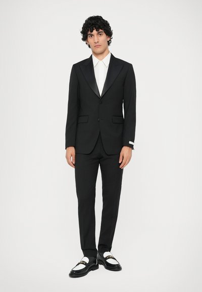 Čierny oblek s kontrastným šálovým klopom, biela dress košeľa, strih prispôsobený postave, a čierno-biele mokasíny s detailmi zlatého hardvéru.