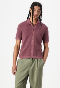 Korte mouwen polo-shirt in stoffig mauve, van een gebreide textuur, met een knoopsluiting aan de voorkant en een zachte kraag, gecombineerd met lichtgroene broek.