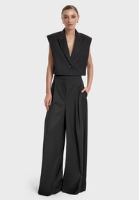 Czarny blazer bez rękawów z guzikiem, w komplecie z spodniami o szerokich nogawkach. Wykonany z teksturowanej tkaniny, strój ma dopasowany krój oraz ostre linie.
