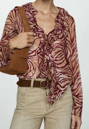 Vrouw draagt een blouse met ruches in bordeauxrood en beige met patroon, beige broek met bruine riem en houdt een bruine suède schoudertas vast.