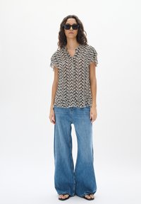 Blusa nera e beige a fantasia con scollo a V, maniche corte a volant, abbinata a jeans a gamba larga blu chiaro e sandali neri.