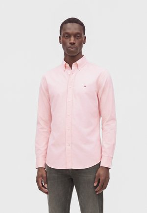 Homme portant une chemise rose pâle à manches longues boutonnée avec un petit logo sur la poitrine et un pantalon gris, debout devant un fond clair uni.
