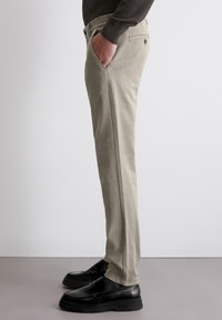 Khakihosen im Slim-Fit-Stil, mit seitlichen Taschen, Gürtelschlaufen und einem strukturierten Stoff. Kombiniert mit schwarzen Dressschuhen.