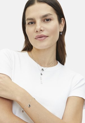 Silberne Schmuckstücke umfassen geschichtete Halsketten mit schwarz-weißen Anhängern und ein passendes Armband mit zwei tropfenförmigen Charms. Das Model trägt ein weißes Hemd.
