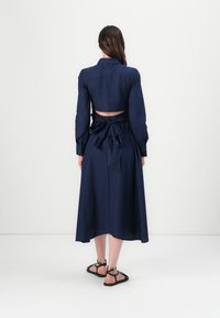 Mörkblå klänning med långa ärmar, kortad baksida och knytning i midjan. Tillverkad av ett mjukt tyg, med en svävande kjol och knappade manschetter.