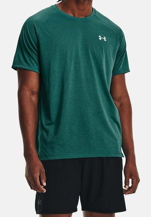 Mann trägt ein grünes kurzärmeliges Sportshirt mit kleinem weißem Logo auf der Brust und schwarze Shorts vor einem einfarbigen Hintergrund.