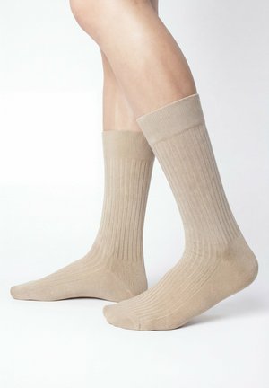 Beine mit beige gerippten Kniestrümpfen stehen auf einer weißen Oberfläche, ein Fuß leicht angehoben.