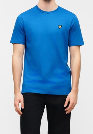 Mann trägt ein schlichtes, leuchtend blaues T-Shirt mit kurzen Ärmeln und kleinem gelbem Vogel-Logo auf der linken Brust sowie schwarze Hose.