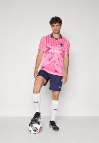 Puma rožinė futbolo marškinėliai su vertikaliomis juostelėmis, tamsiai mėlyni šortai, juodi sportbačiai ir balti kojinės. Žaidėjas stovi ant raštuoto futbolo kamuolio.
