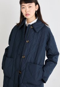 Veste matelassée marine avec un grand col, deux poches avant et des boutons marron. Elle présente une texture lisse et un motif de couture en diamant.