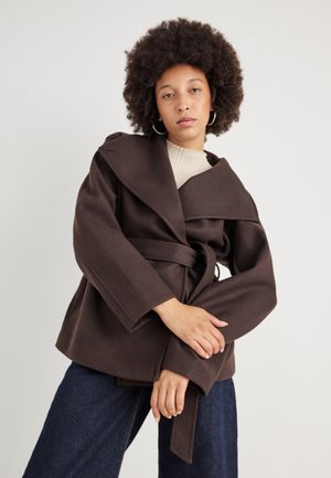 Brun uldblandet omslagsfrakke med brede revers, bælte i taljen og klokkeærmer. Båret over en ribbet beige top, parret med mørk denim.