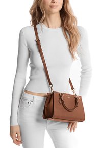 Bolso sintético marrón con herrajes en tono dorado y dobles asas superiores. Presenta una correa de hombro desmontable. El modelo lleva una blusa blanca de canalé y jeans blancos.