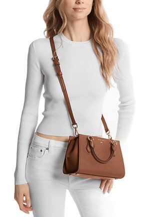 Bolso sintético marrón con herrajes en tono dorado y dobles asas superiores. Presenta una correa de hombro desmontable. El modelo lleva una blusa blanca de canalé y jeans blancos.