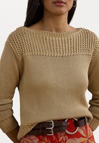 Beige strikkegenser med teksturert overdel og ribbestrikkede mansjetter, kombinert med et brunt dobbelbelte med en sirkulær metallspenne.