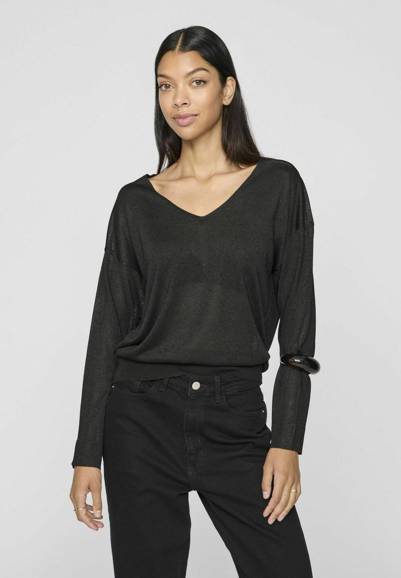 Musta, pitkähihainen neulepaito, jossa on V-kaula-aukko, rento malli ja hieman läpinäkyvä rakenne, yhdistettynä mustiin korkeavyötäröisiin housuihin.