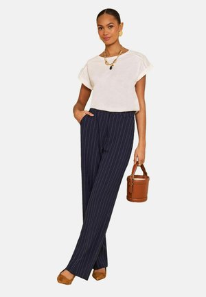 Žena nosi bielu blúzku s krátkym rukávom, námornícky modré široké nohavice s jemným prúžkom, hnedé špicaté topánky, zlaté šperky a drží hnedú koženú kabelku vo forme vedierka.