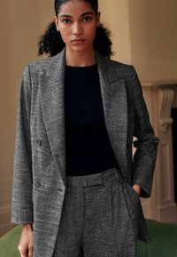 Grå blazer med et struktureret mønster, dobbeltknapsdesign og store reverser, parret med en mørk ribbet top og matchende bukser.