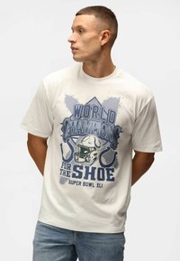 Kortærmet cremet t-shirt med en blå grafisk design, der inkluderer tekst og en fodboldhjelm, fremstillet af bomuldsstof med en glat tekstur.