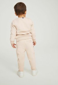 Lys rosa sweatshirt og matchende sweatpants, blød tekstur, tætsiddende manchetter, subtil logo-detail på bukserne, båret med hvide sneakers.