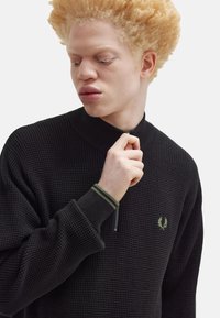 Maglione zip-up in tessuto waffle nero con accenti verdi nel colletto e nei polsini; presenta un piccolo logo ricamato sul petto.
