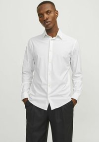 Camicia bianca a maniche lunghe in cotone liscio, con colletto classico, chiusura con bottoni davanti e vestibilità su misura. Abbinata a pantaloni scuri.