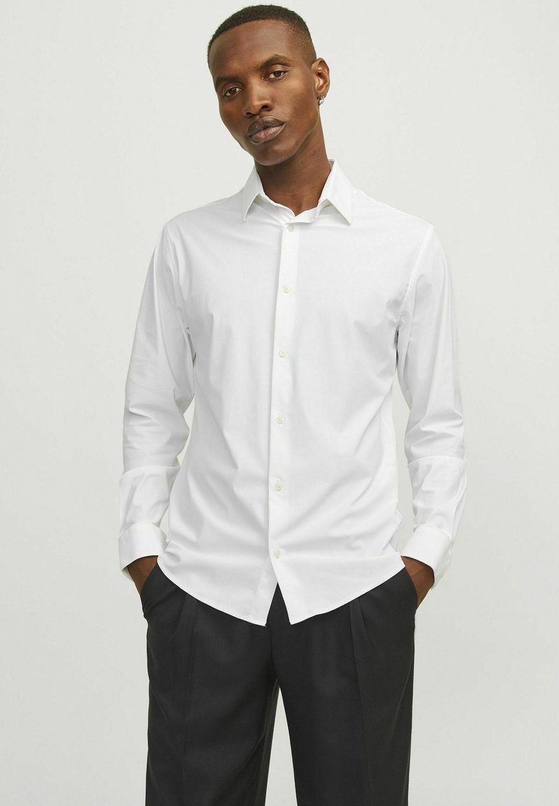Camicia bianca a maniche lunghe in cotone liscio, con colletto classico, chiusura con bottoni davanti e vestibilità su misura. Abbinata a pantaloni scuri.