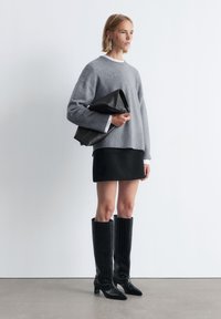 Grauer Strickpullover mit kastigem Schnitt, kombiniert mit einem schwarzen Minirock. Schwarze kniehohe Stiefel aus Krokoledertextur. Hält eine schwarze Clutch-Tasche.