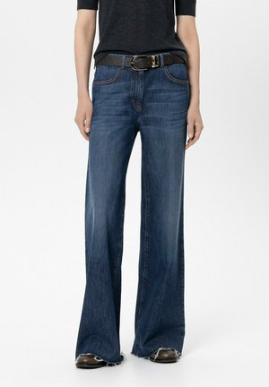 Person wearing dark blue wide-leg jeans with frayed hems, a black belt, and dark shoes against a plain white background.