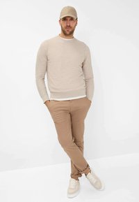 Beige Strickpullover mit einem hellen Kragen, kombiniert mit hellbraunen Hosen. Das Model trägt eine passende beige Mütze und cremefarbene Sneaker mit braunen Akzenten.