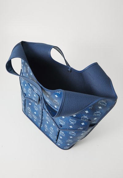 Μπλε τσάντα tote MCM με λευκό μονογράμμα, δύο μπροστινές τσέπες και ανοιχτή κορυφή, εμφανίζεται από γωνιακή επάνω προοπτική σε λευκό φόντο.