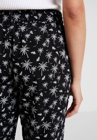 Pantalons noirs avec motif de palmiers blancs, dotés d'une taille élastique et d'une texture de tissu douce. La vue arrière montre les détails du motif de palmiers.